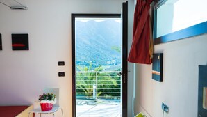 A room with a bed, a small table with a red pot of flowers, a chair, and a sliding glass door leading to a balcony with a mountain view. - Natur Hotel Tanca (Cardedu)