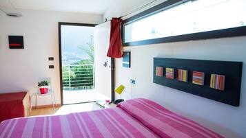Un dormitorio con una cama, una silla, una mesita con un balde rojo y una ventana con vistas al verdor.