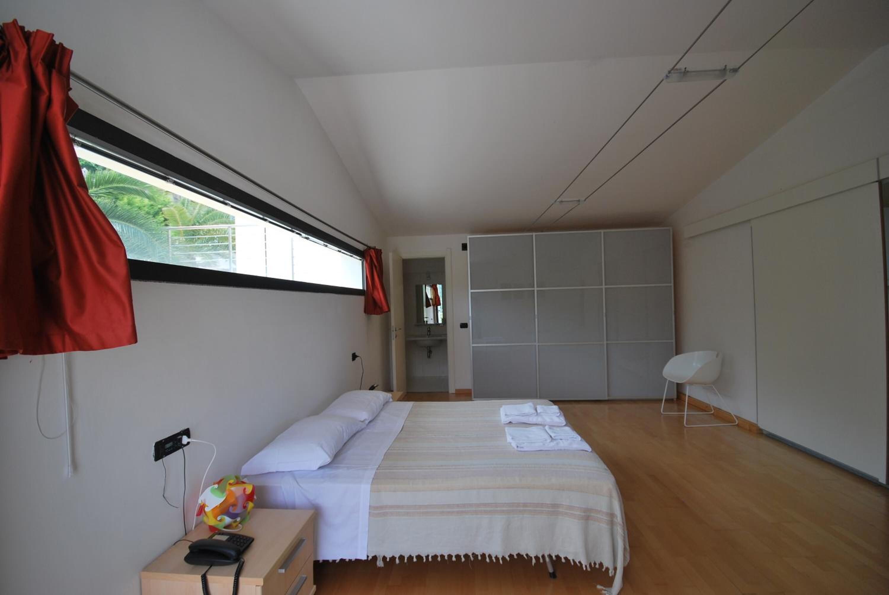 Um quarto moderno com uma cama grande, uma mesinha de cabeceira com um telefone e um objeto colorido, uma janela com cortinas vermelhas e vista para o verde lá fora.