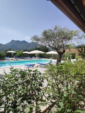 Un espace piscine doté de chaises longues et de parasols, entouré de verdure et avec des montagnes en toile de fond.