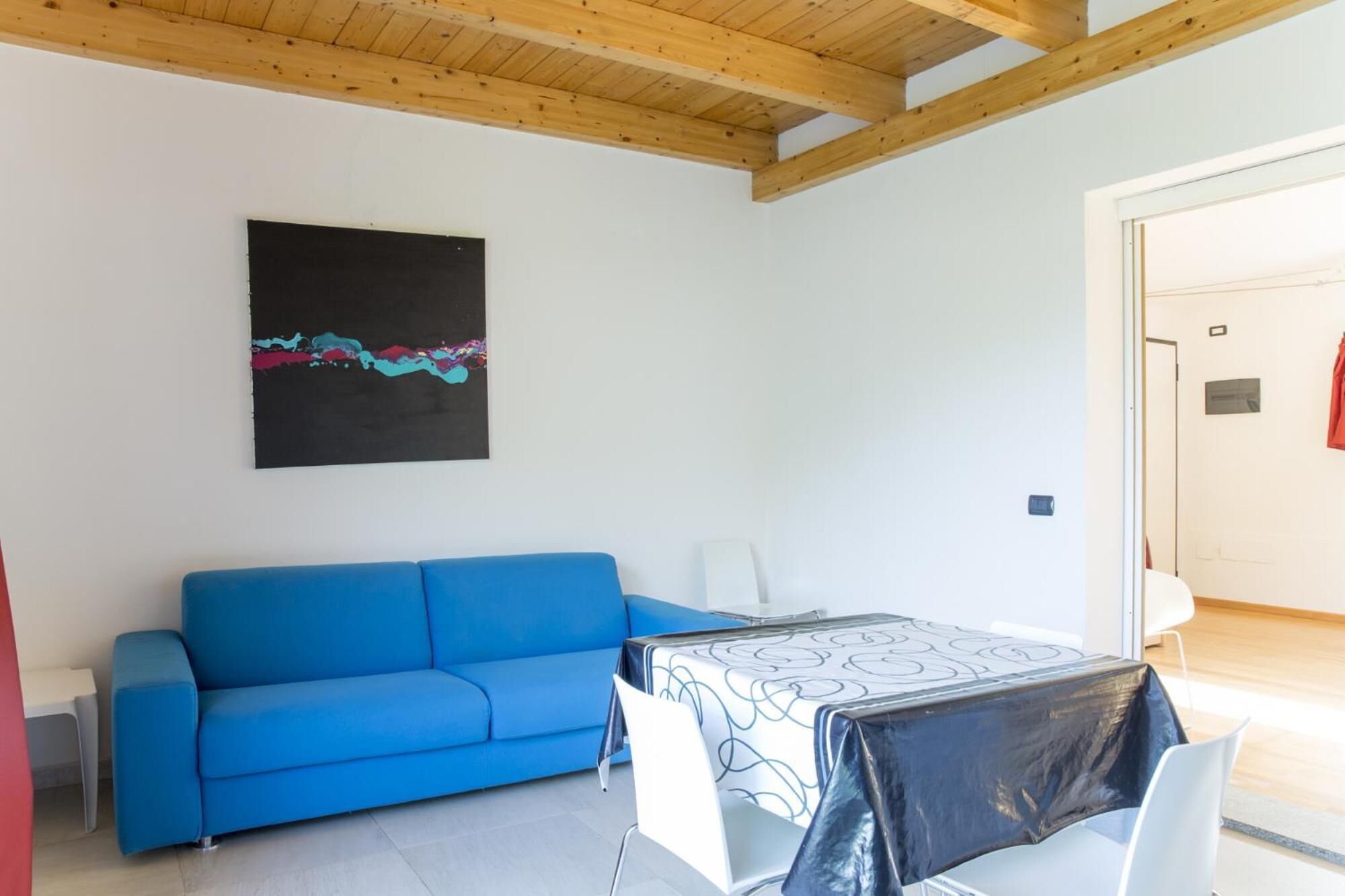 Une pièce avec un canapé bleu, une table recouverte d’une nappe à motifs et un tableau abstrait noir accroché au mur.