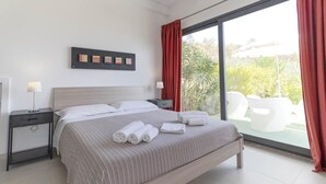 A modern hotel room with a large bed, a nightstand, a wall-mounted artwork, and a view of outdoor seating. - Natur Hotel Tanca (Cardedu)