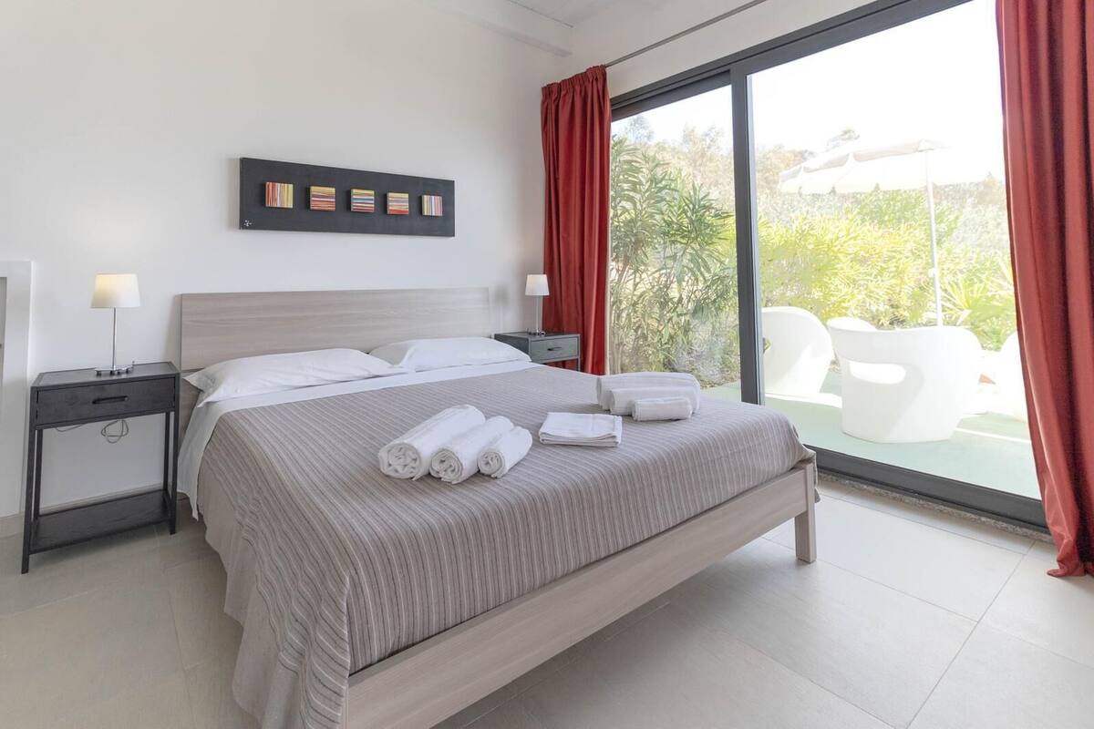 A modern hotel room with a large bed, a nightstand, a wall-mounted artwork, and a view of outdoor seating.