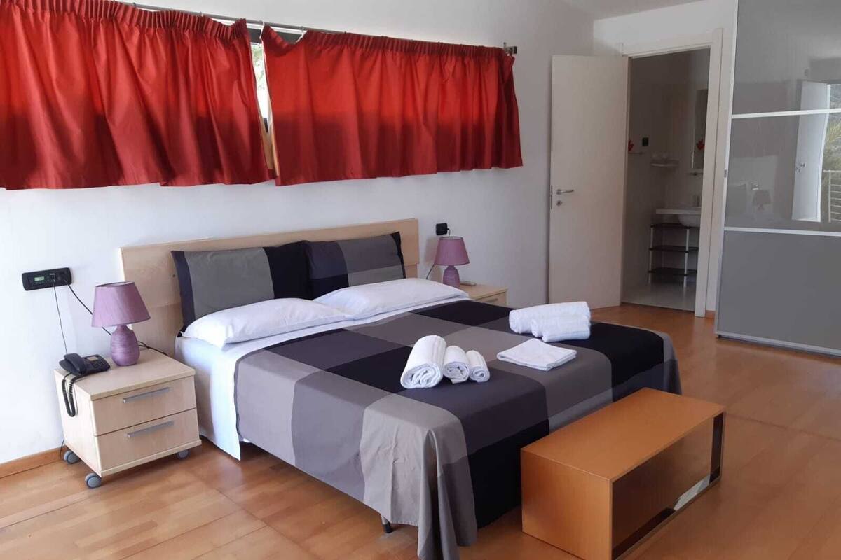 A neatly made bed with a grey and black bedspread, two pillows, and rolled towels on a wooden floor.