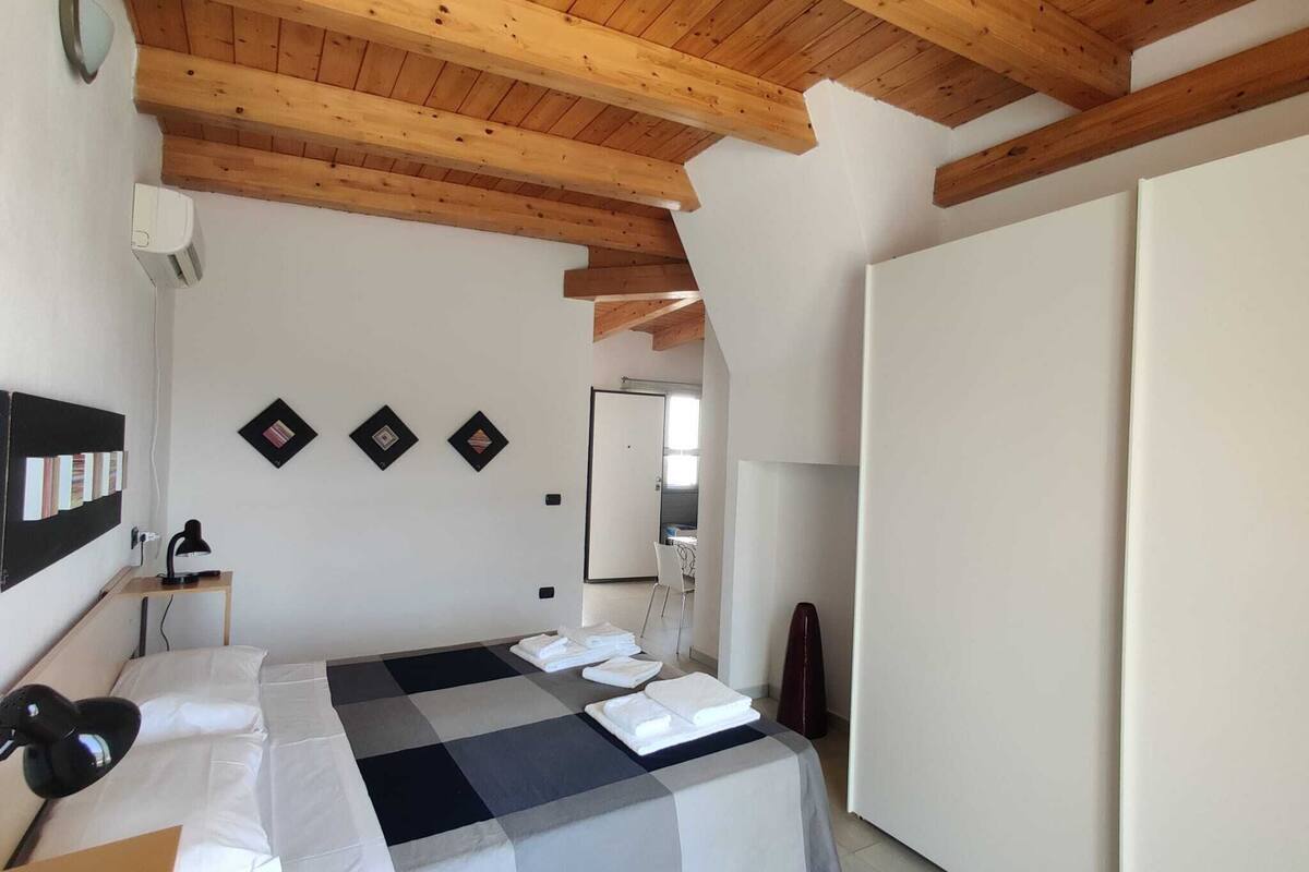 A bedroom with a checkered bedspread, wooden ceiling beams, a desk with a lamp, and a wall-mounted shelf with books.