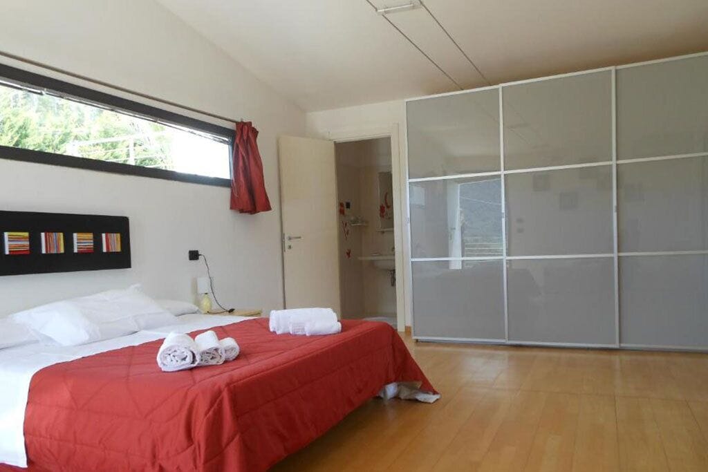 A hotel room with a bed, a wardrobe, a window with curtains, and a wall-mounted shelf with items.