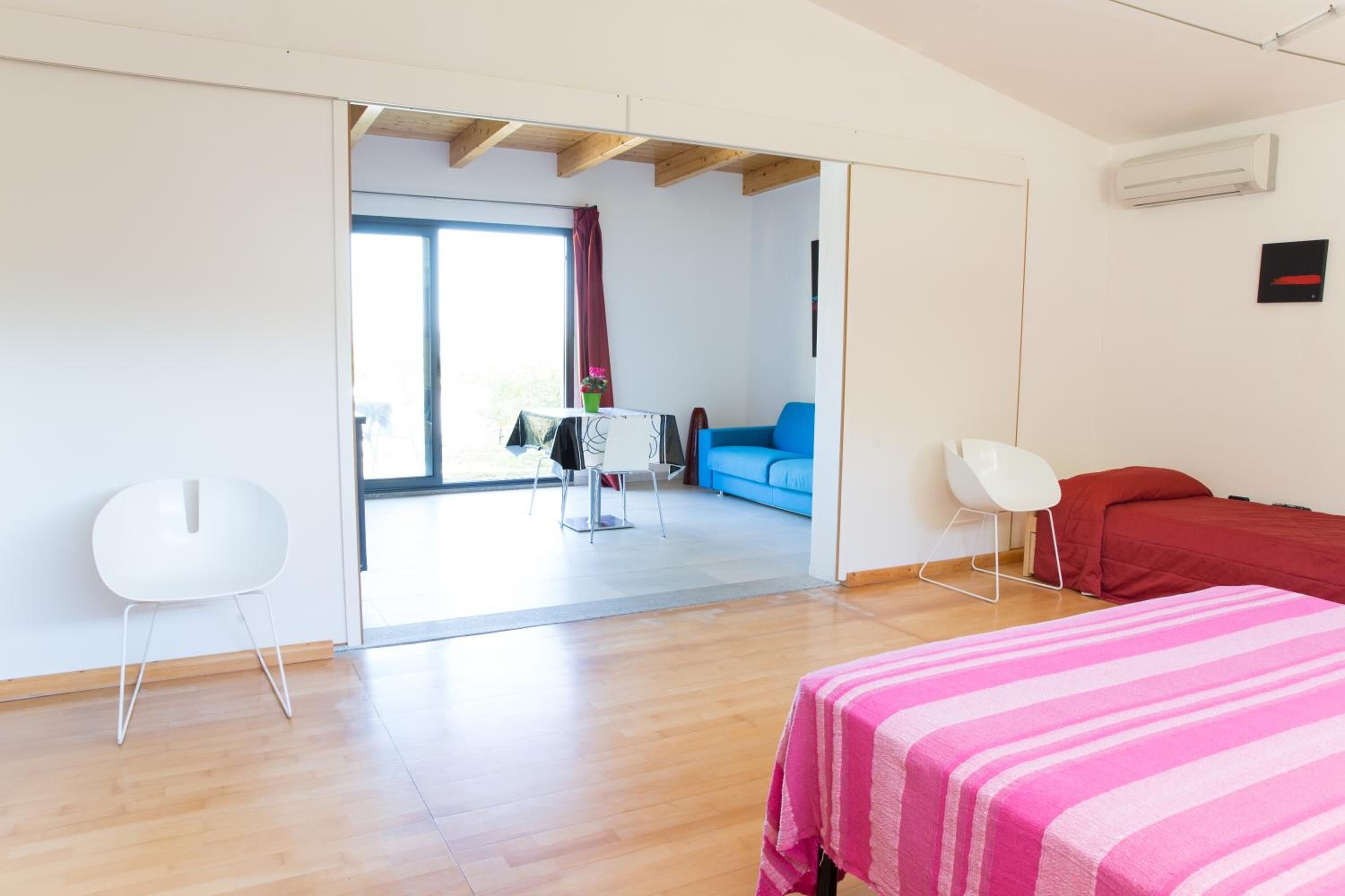 A modern room with a wooden floor, a bed with a pink striped bedspread, a white chair, a blue sofa, and a table with a vase of flowers.