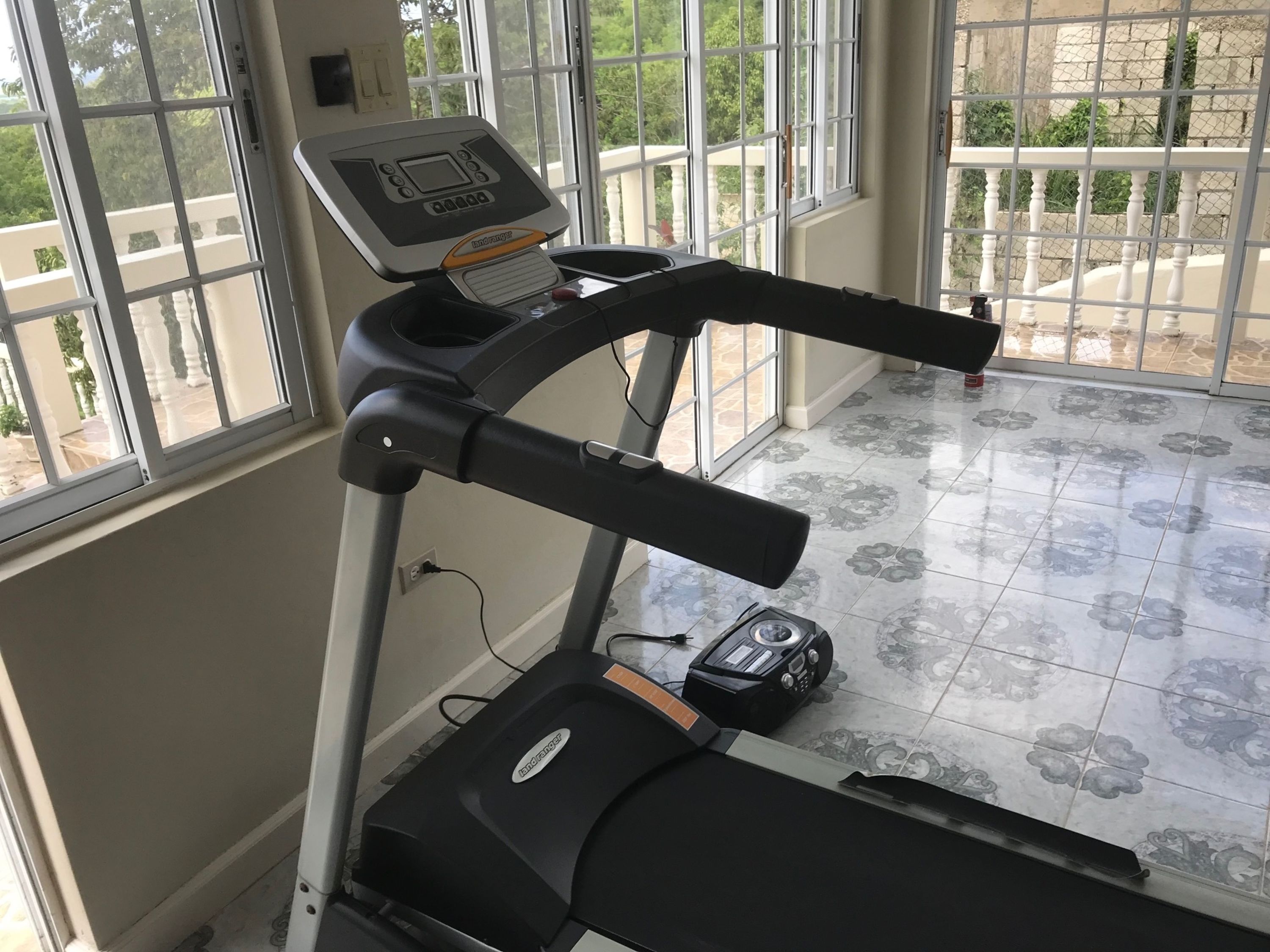 A treadmill in a room with large windows, a patterned floor, and a visible electrical outlet.