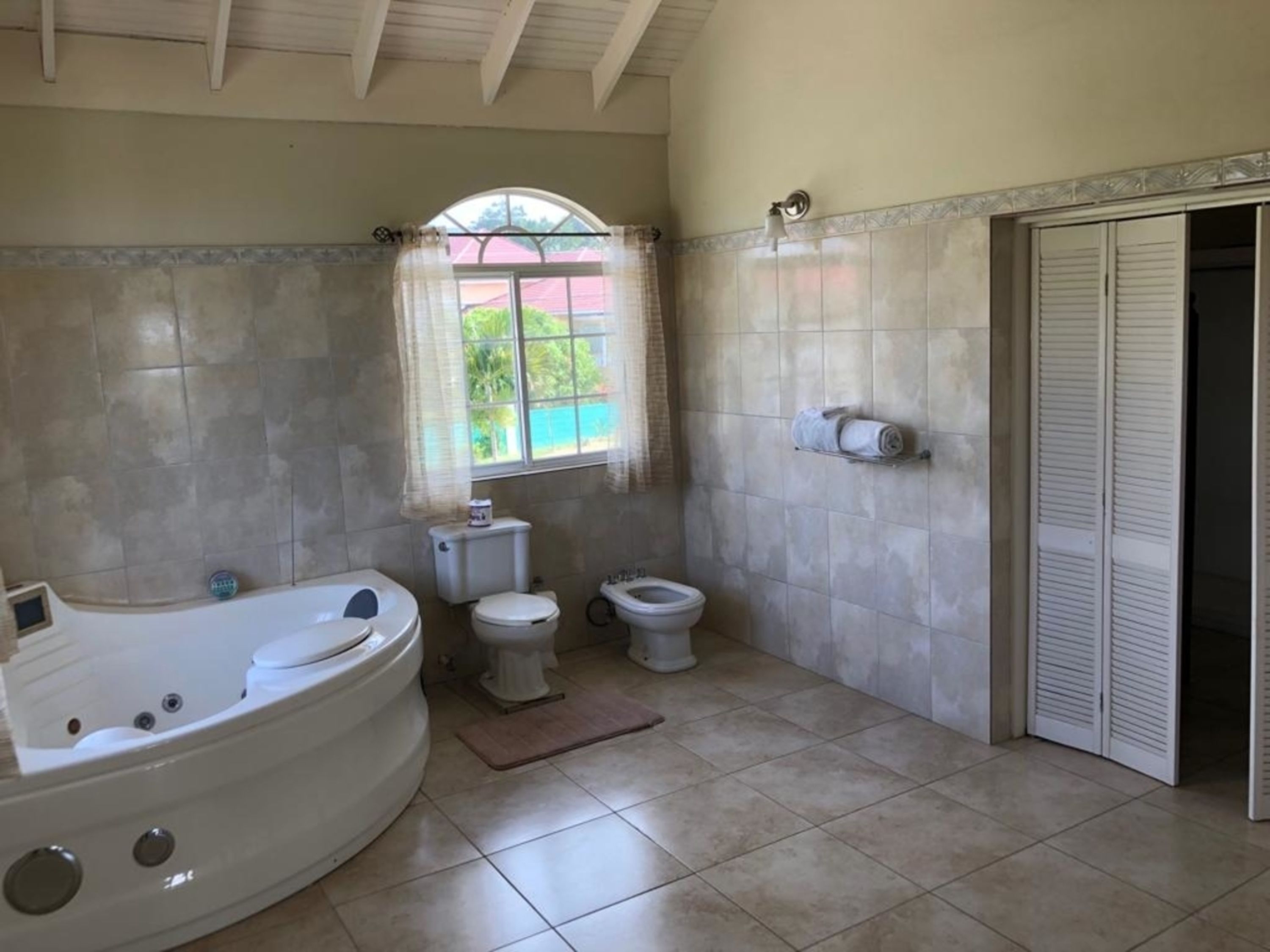 Une salle de bain avec une baignoire, deux toilettes, une fenêtre avec des rideaux et un sol carrelé.