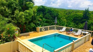 A rectangular pool with a white fence and a white balustrade, surrounded by lounge chairs and umbrellas, with lush greenery in the background.