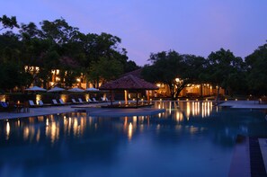 Un espace piscine paisible au crépuscule, avec des chaises longues et des parasols, entouré d’arbres et d’un bâtiment dont l’éclairage est chaleureux.
