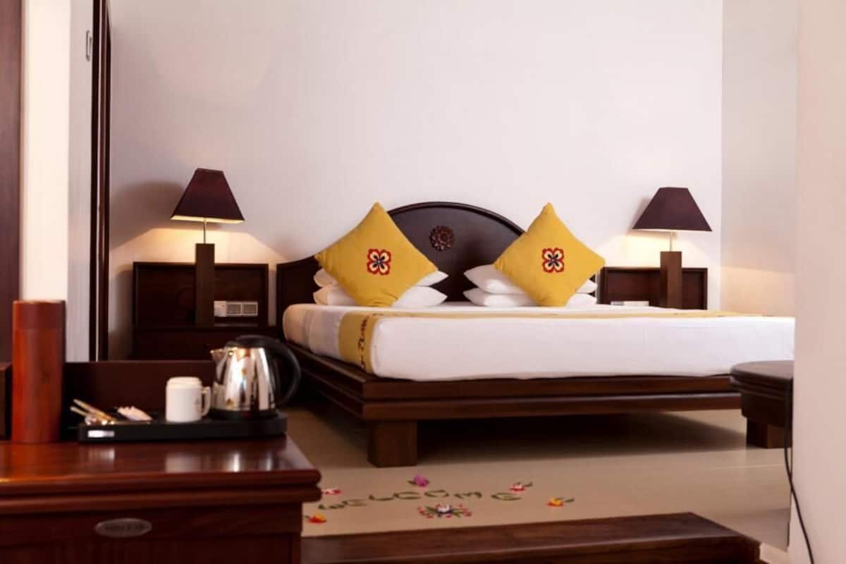 Quarto de hotel com cama, duas luminárias de cabeceira, mesa de cabeceira com máquina de café e tapete de boas-vindas.