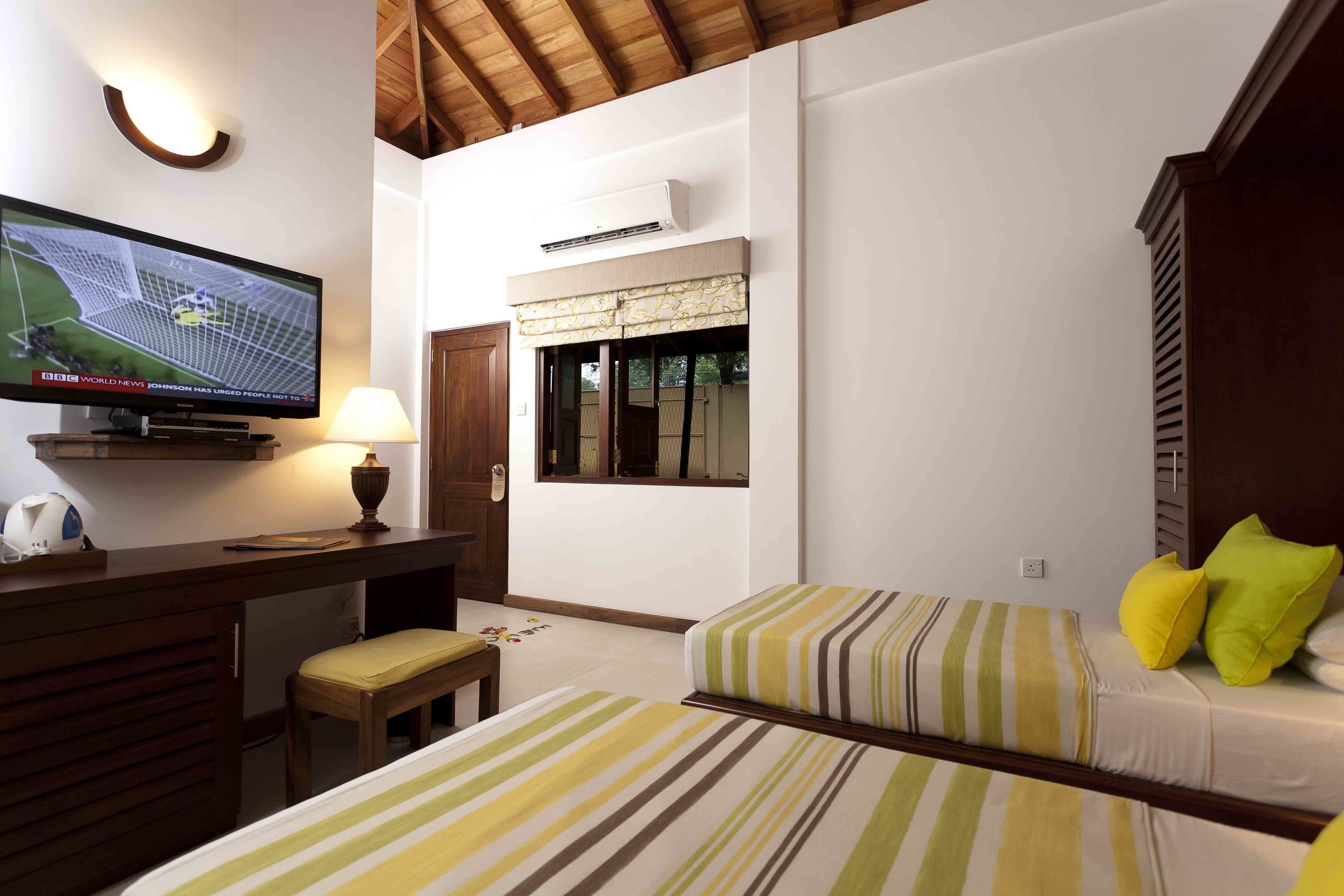 A hotel room with a television, a desk, two beds with striped bedding, a window with blinds, and an air conditioning unit.