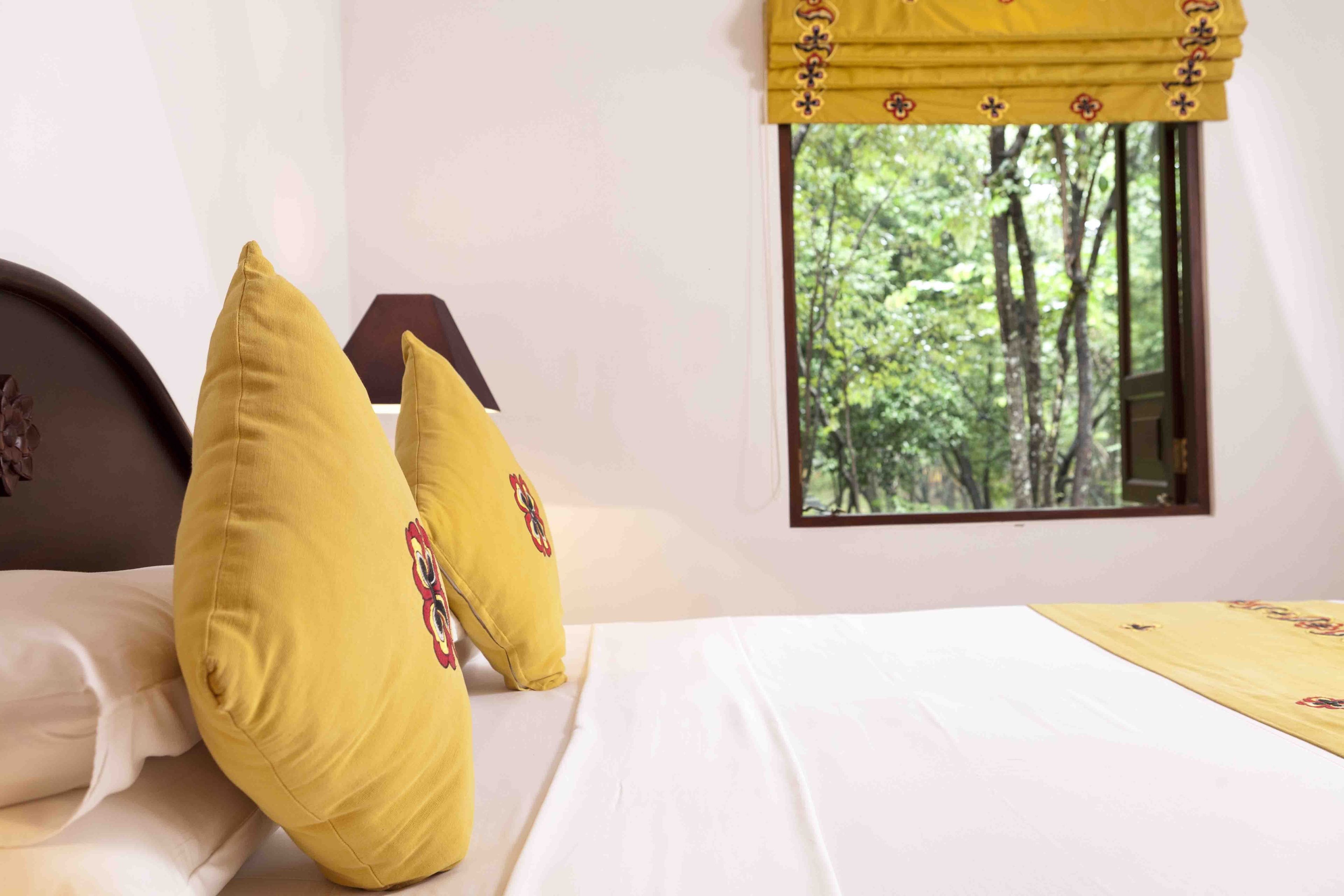 A bed with white linens and two yellow pillows, a window with a view of trees, and a yellow curtain with a floral pattern.