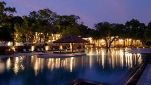A swimming pool with lounge chairs and umbrellas, surrounded by trees and a building with lights. - Amaya Lake Dambulla (Kandalama)