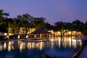 Una piscina con sillas de descanso y sombrillas, rodeada de árboles y un edificio con luces.