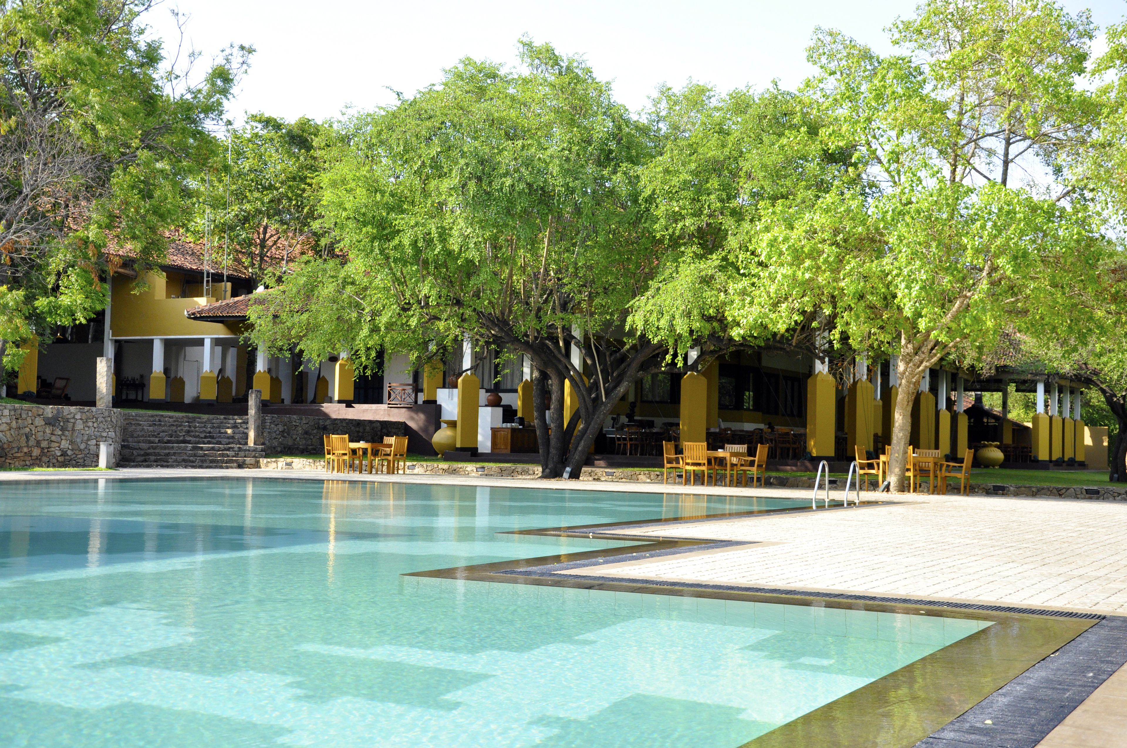 Una piscina con agua cristalina, rodeada de edificios blancos y amarillos, y árboles que dan sombra.