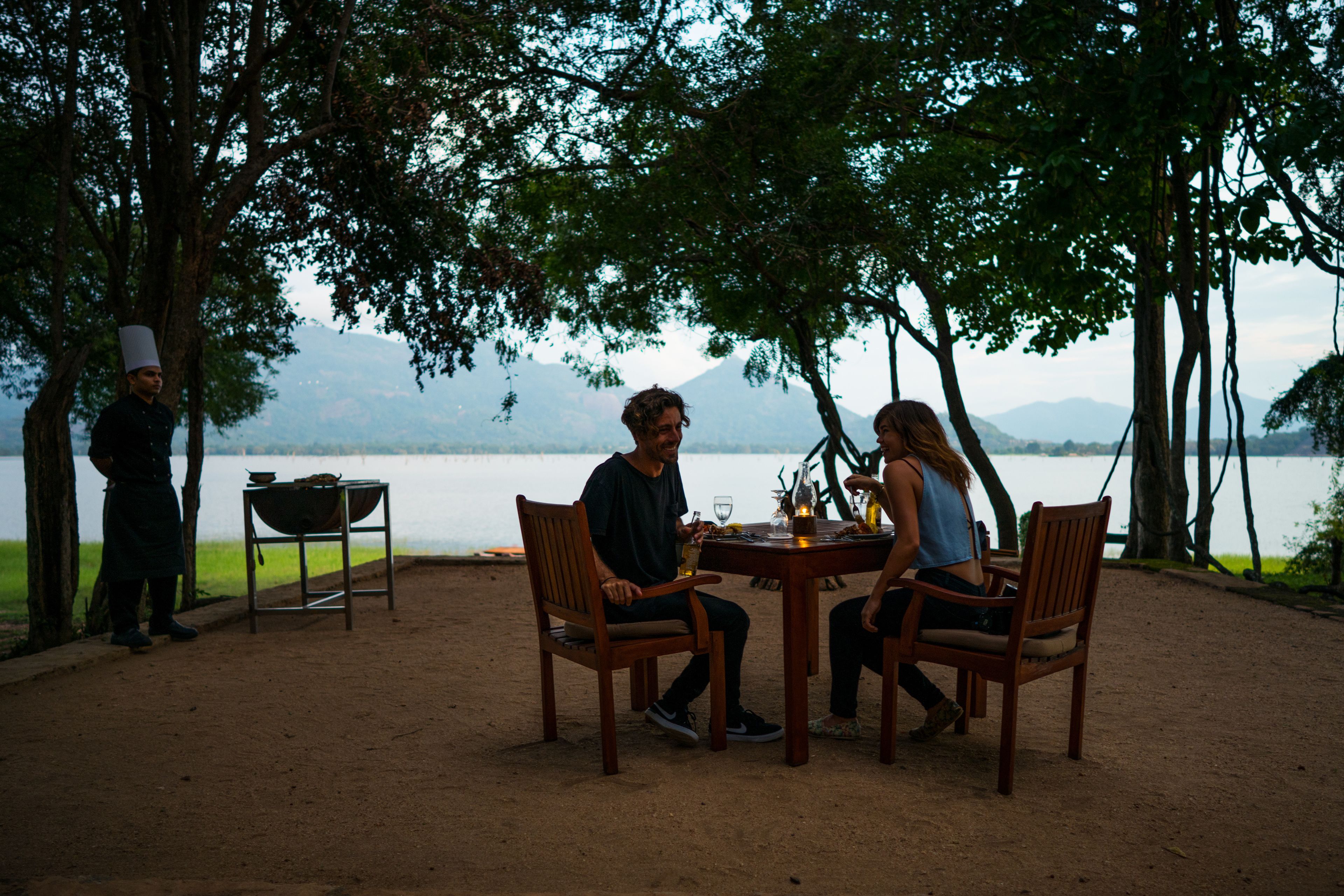 A dining area with a table and chairs, a person standing by a grill, and a view of a lake with mountains in the background.