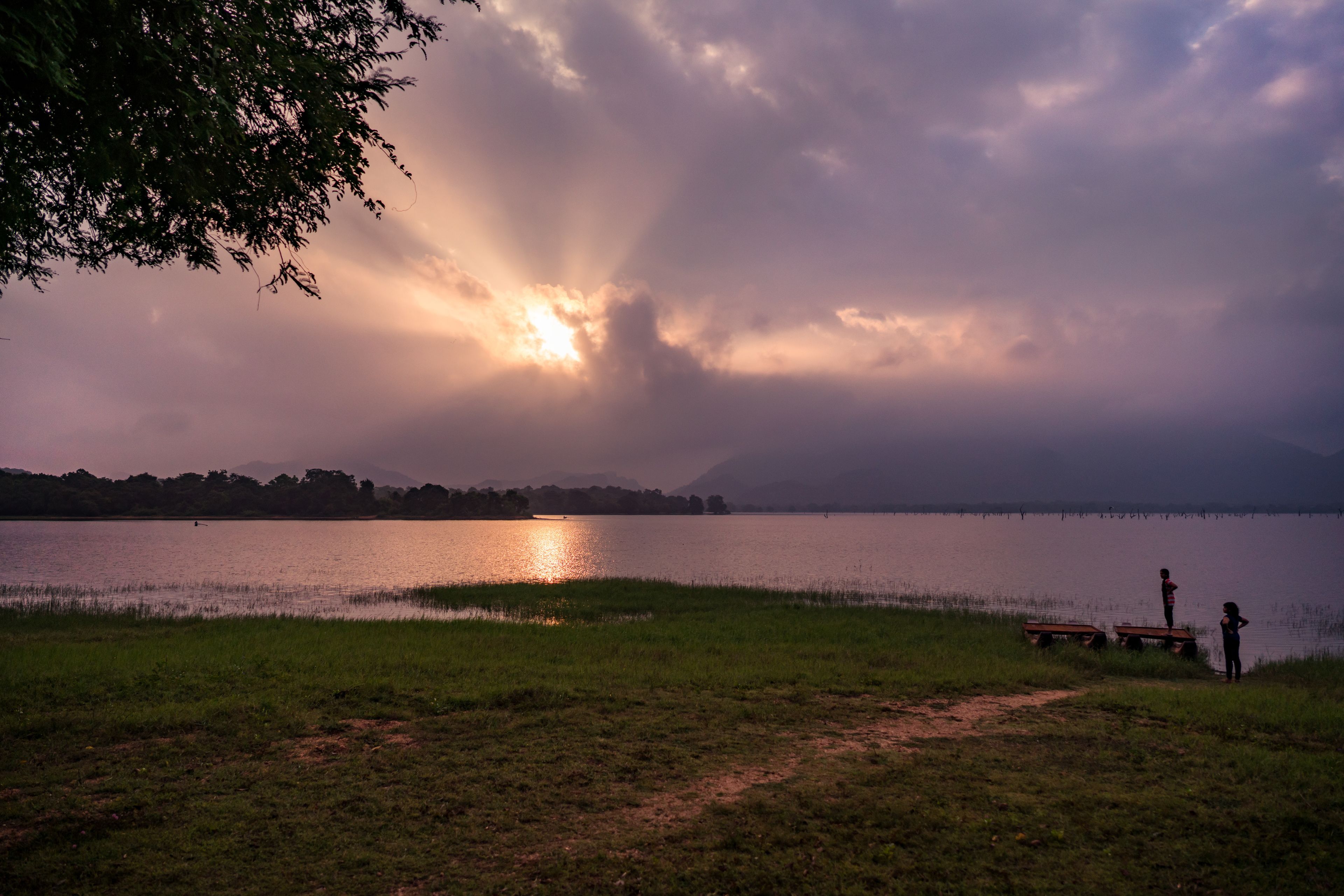 A lakeside view with a path leading to a dock, a bench, and two people standing by the water.
