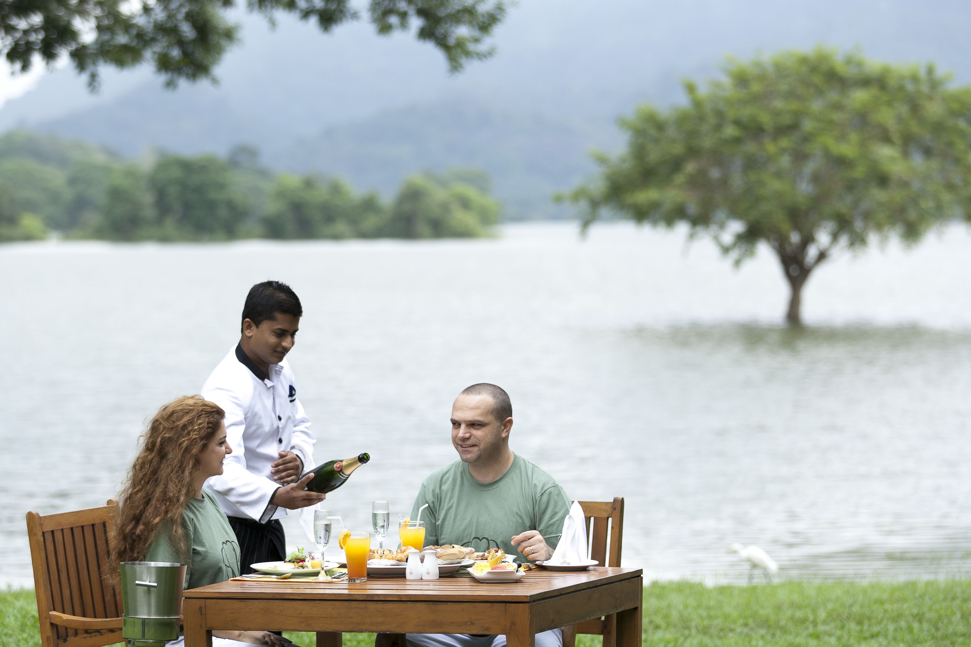 A dining table with food and drinks, a waiter, and a lake view.