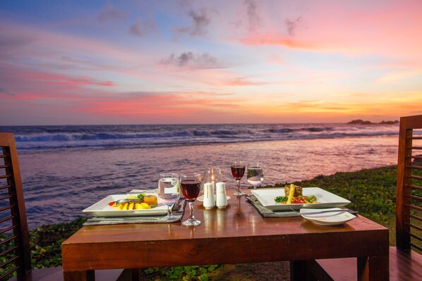 A table set with plates of food and glasses of wine, overlooking the ocean at sunset.