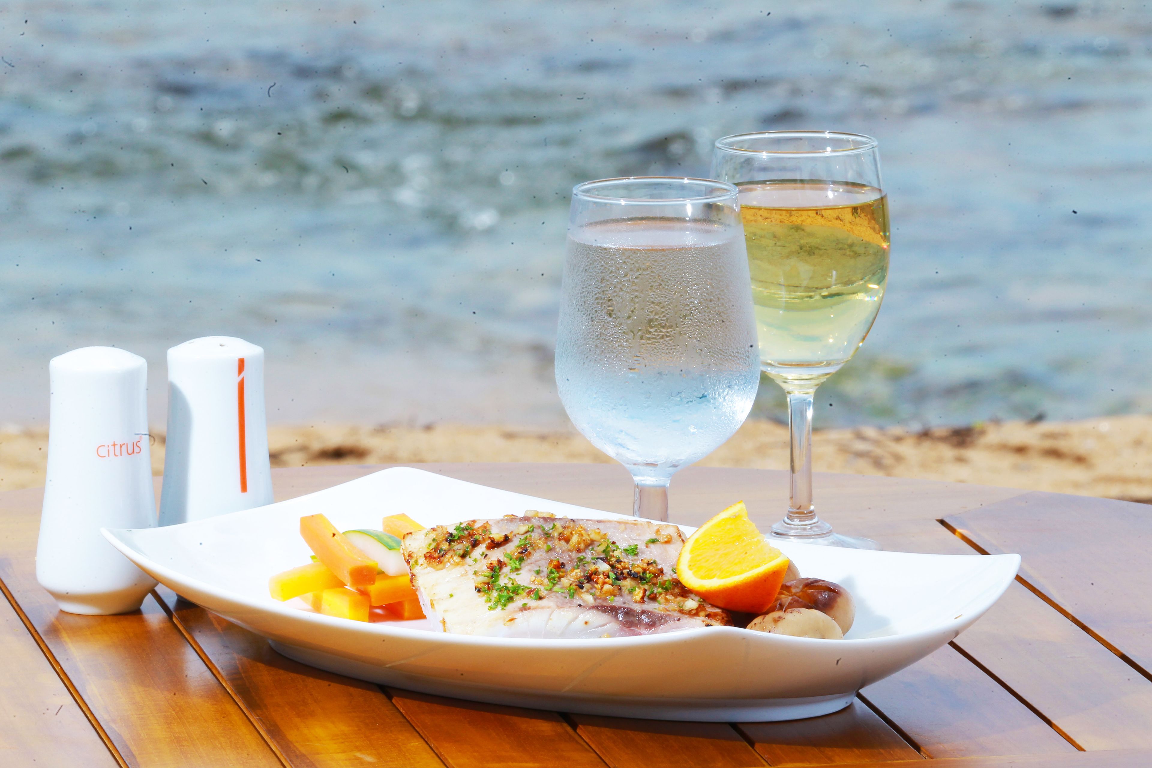 A seafood dish on a white plate with two glasses of white wine and a slice of orange on a wooden table by the sea.