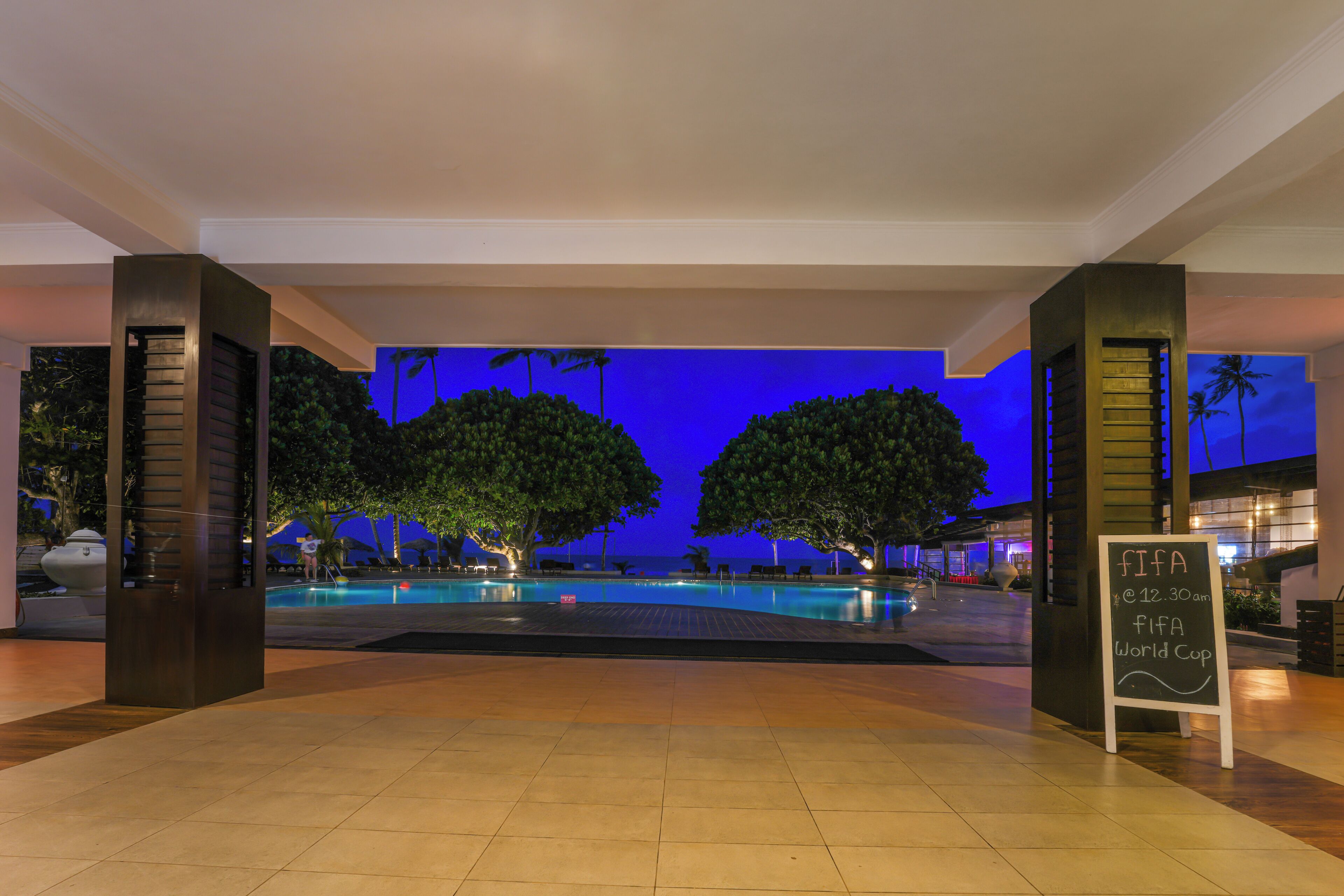 Un espace piscine avec vue sur l’océan au crépuscule, un panneau indiquant l’heure du début de la Coupe du monde de la FIFA, et un sol carrelé.