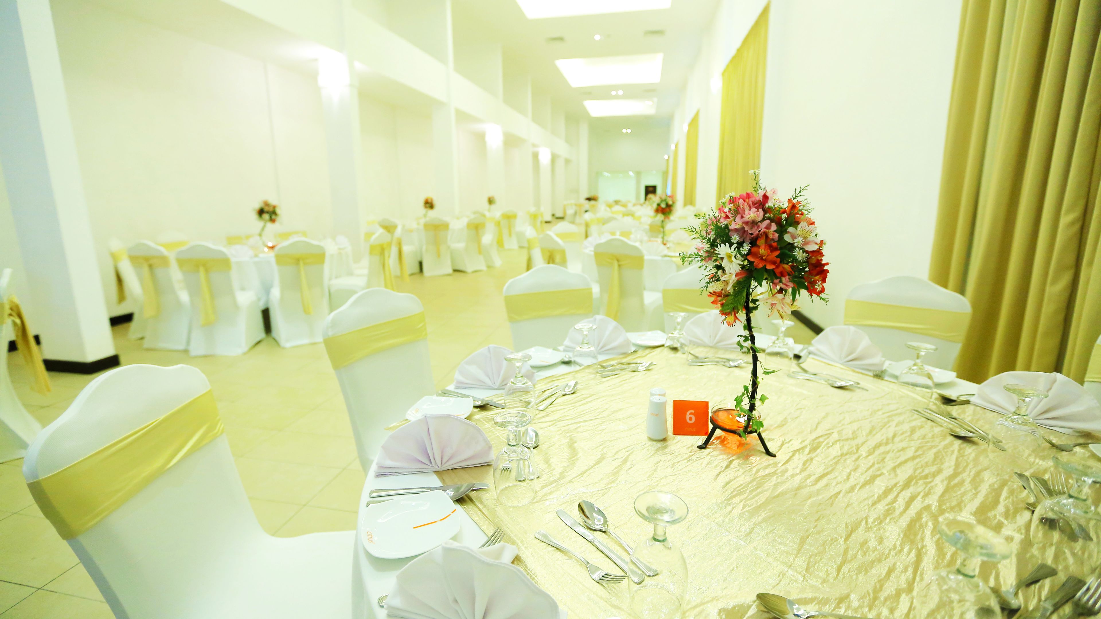 Un salón de banquetes con mesas redondas dispuestas para un evento formal, con sillas blancas adornadas con cintas amarillas, un arreglo floral central y tarjetas de lugar numeradas.