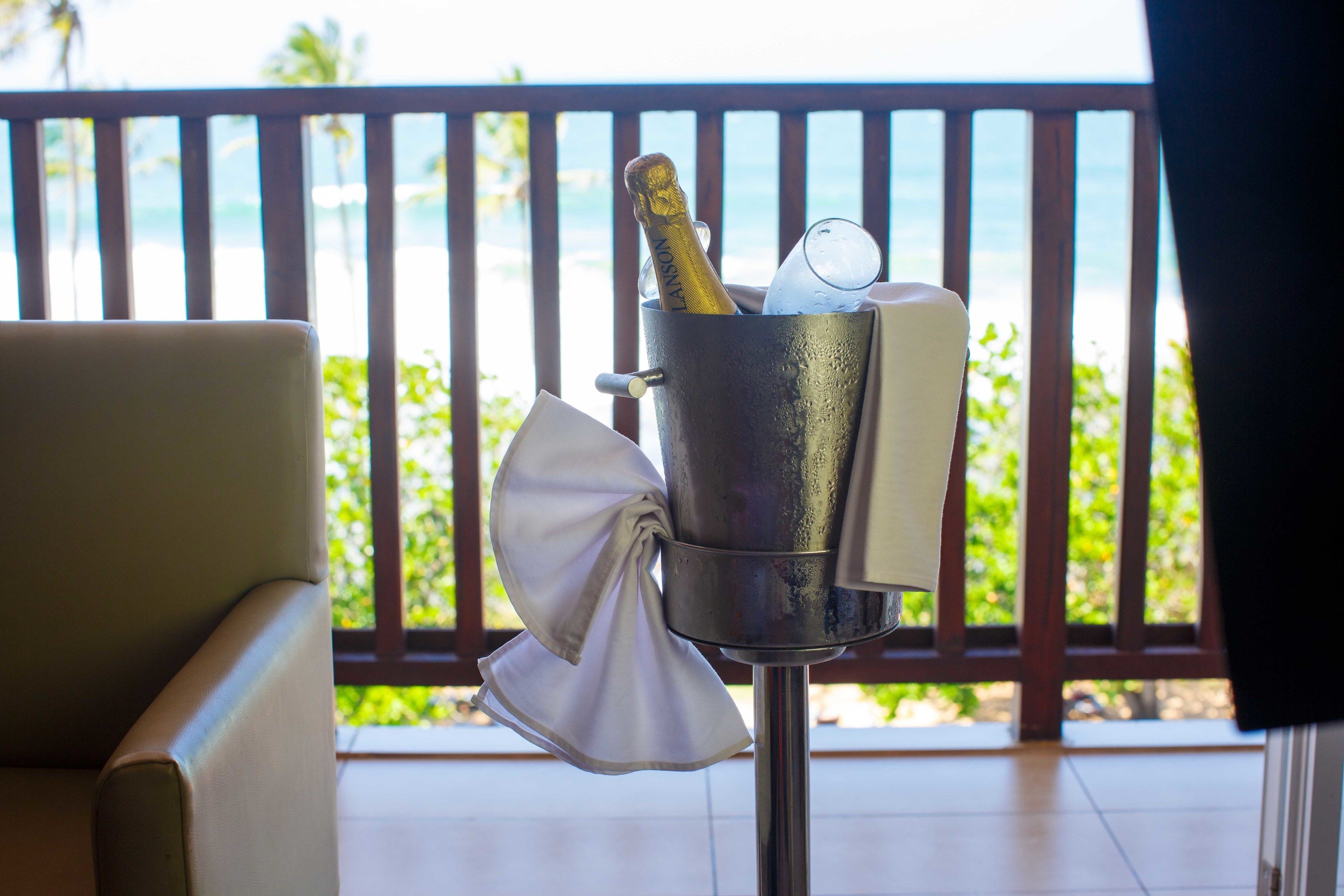 Een metalen emmer met een fles champagne, een glas en een servetje op een balkon met houten leuning en uitzicht op de oceaan.