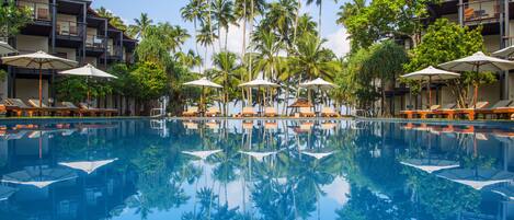 A swimming pool with lounge chairs and umbrellas, surrounded by palm trees and hotel buildings.