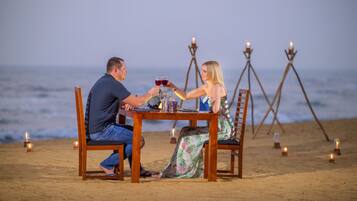 Una pareja cenando en una mesa de madera en la playa, con velas y vista al océano.