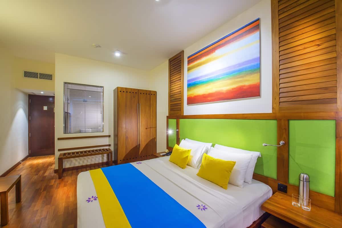 A hotel room with a bed, wooden wardrobe, a bench, a window with blinds, and a colorful abstract painting on the wall.