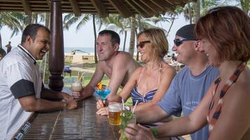 A group of people sitting at an outdoor table with drinks, palm trees, and a beach in the background.