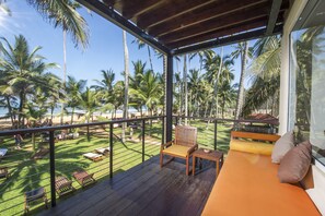 Uma área externa coberta com deck de madeira, espaço de estar acolchoado e vista para uma praia com palmeiras.
