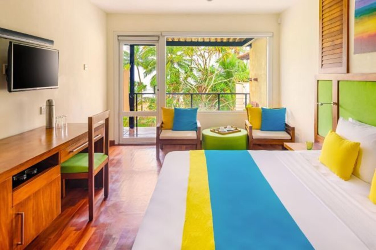 A hotel room with a bed, a desk, a chair, a sofa, and a balcony with a view of palm trees.