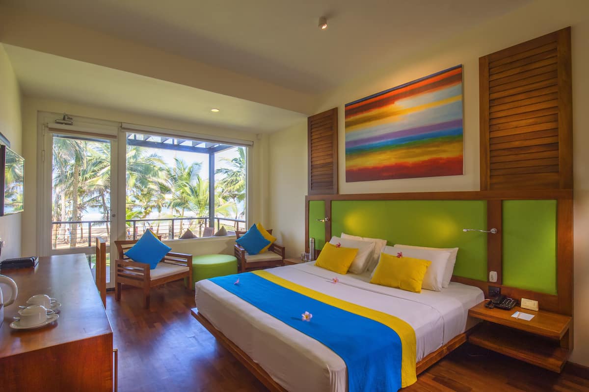 A hotel room with a large bed, a desk with a chair, a window with a view of palm trees, and a colorful painting on the wall.