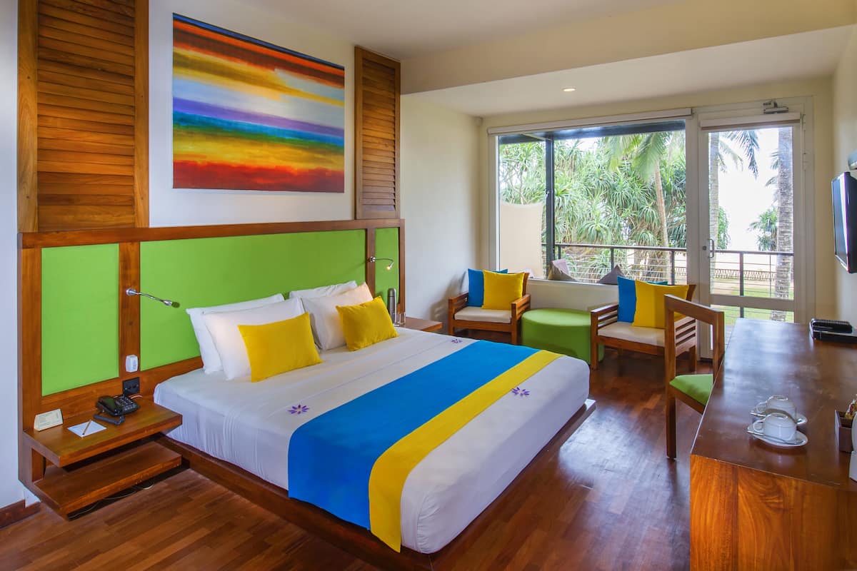 A hotel room with a large bed, a green accent wall, a colorful painting, and a balcony with a view of greenery.