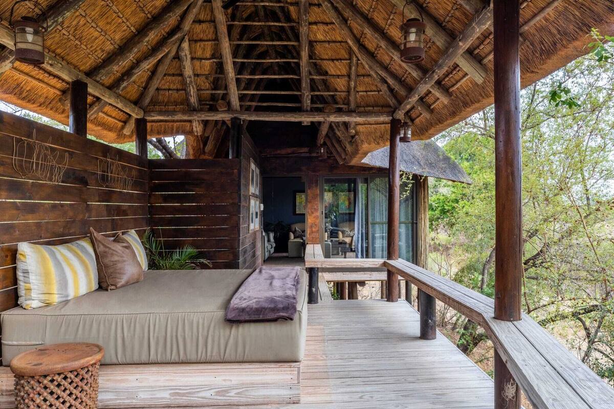 A wooden deck with a thatched roof, a sofa with cushions, and a view of the surrounding forest.