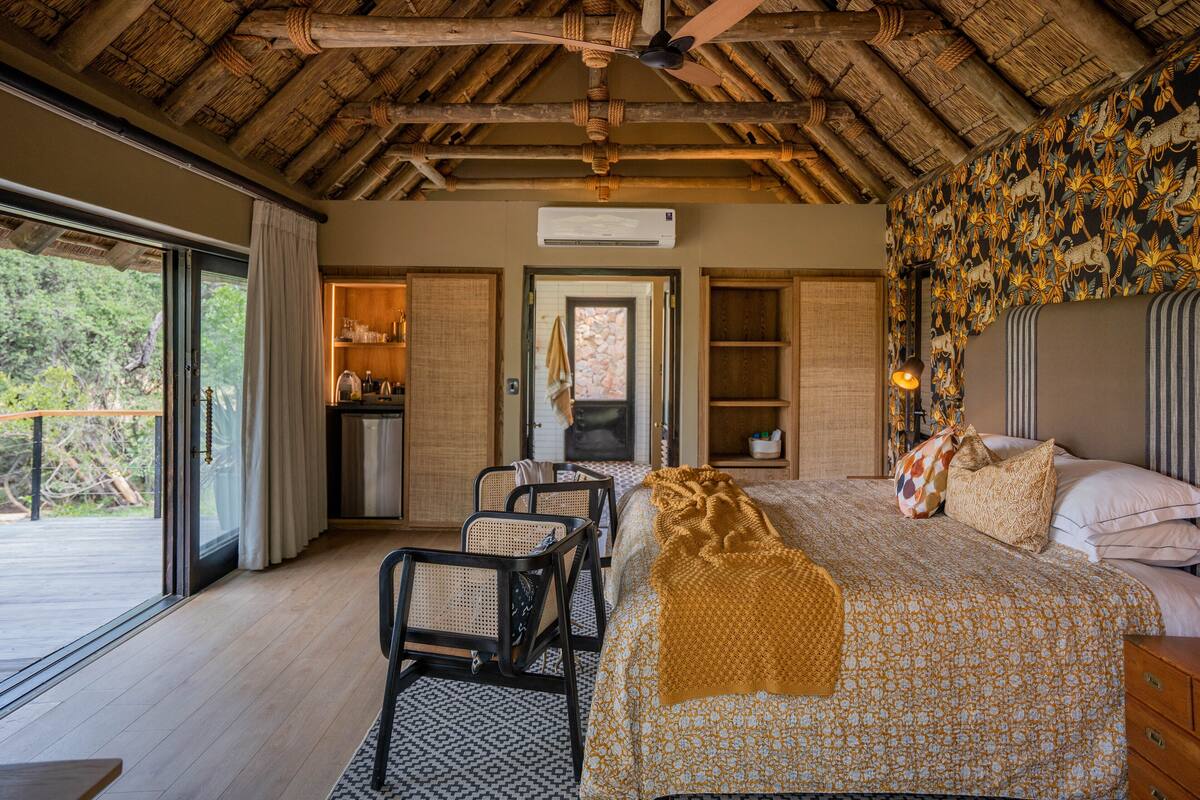 A bedroom with a bed, a desk, and chairs, a ceiling fan, and a view of the outdoors.