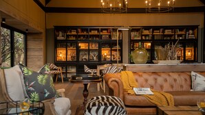 Un salon chaleureux avec un canapé en cuir, des fauteuils en osier et une statue de zèbre. Une bibliothèque remplie de livres et d’objets décoratifs se trouve au fond de la pièce.