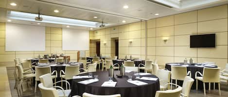 Un salón de conferencias con varias mesas y sillas, equipado con proyectores y pantallas, en un entorno moderno y bien iluminado de un hotel.