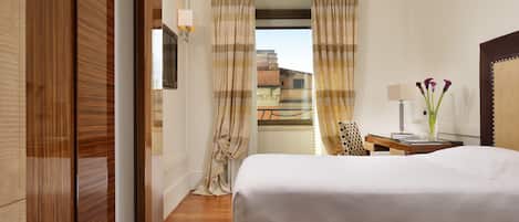A hotel room with a bed, a desk with a lamp, a chair, and a view of buildings through the curtains.