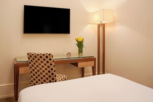 A hotel room with a bed, a desk with a chair, a TV mounted on the wall, and a lamp.