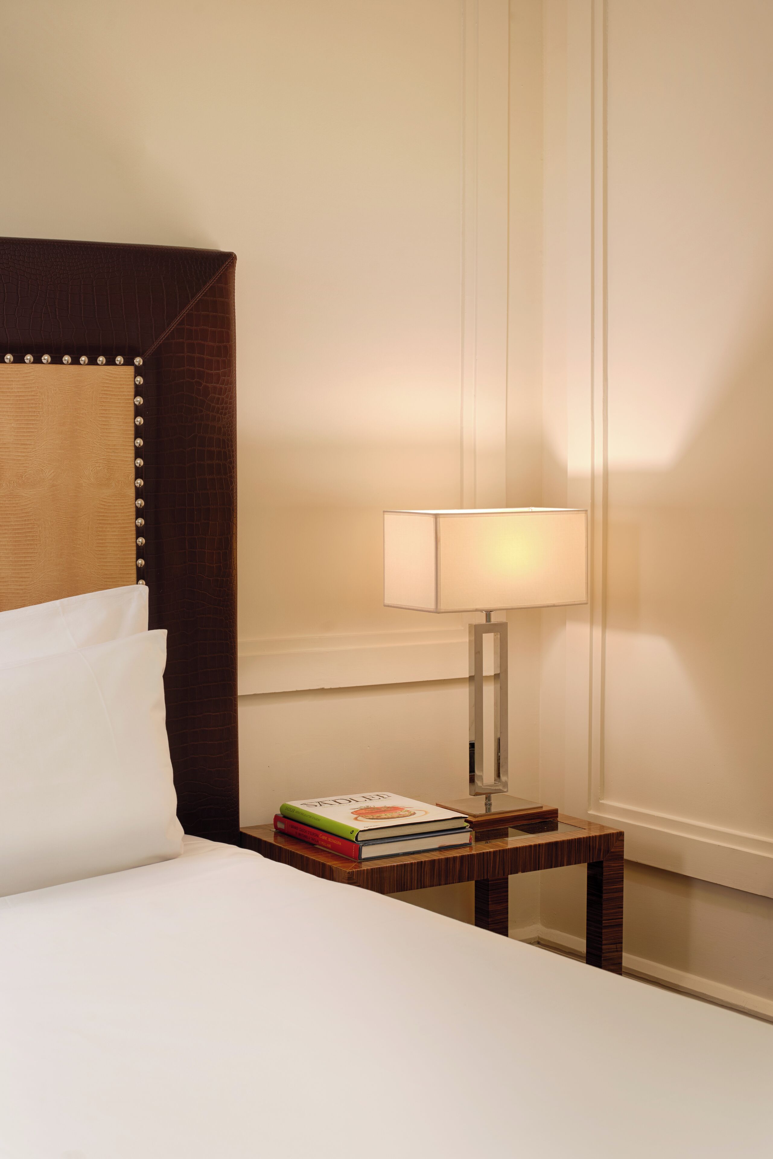 A neatly made bed with white linens, a wooden bedside table with a lamp, and a stack of books.