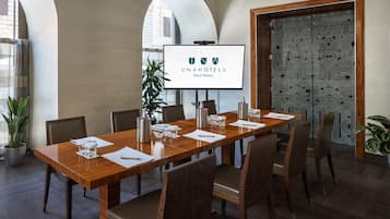 Een vergaderzaal met een lange houten tafel, stoelen, een projectiescherm waarop 'UNAHOTELS Decò Roma' staat, en een deur met een patroon.