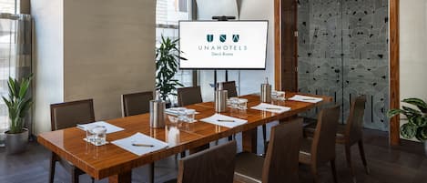 A conference room with a long wooden table, chairs, a projector screen displaying 'UNAHOTELS Decò Roma,' and a patterned door.
