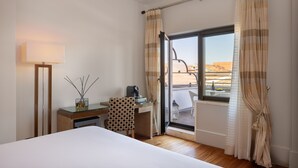 A hotel room with a bed, a desk with a lamp, a chair, and a view of the outside.