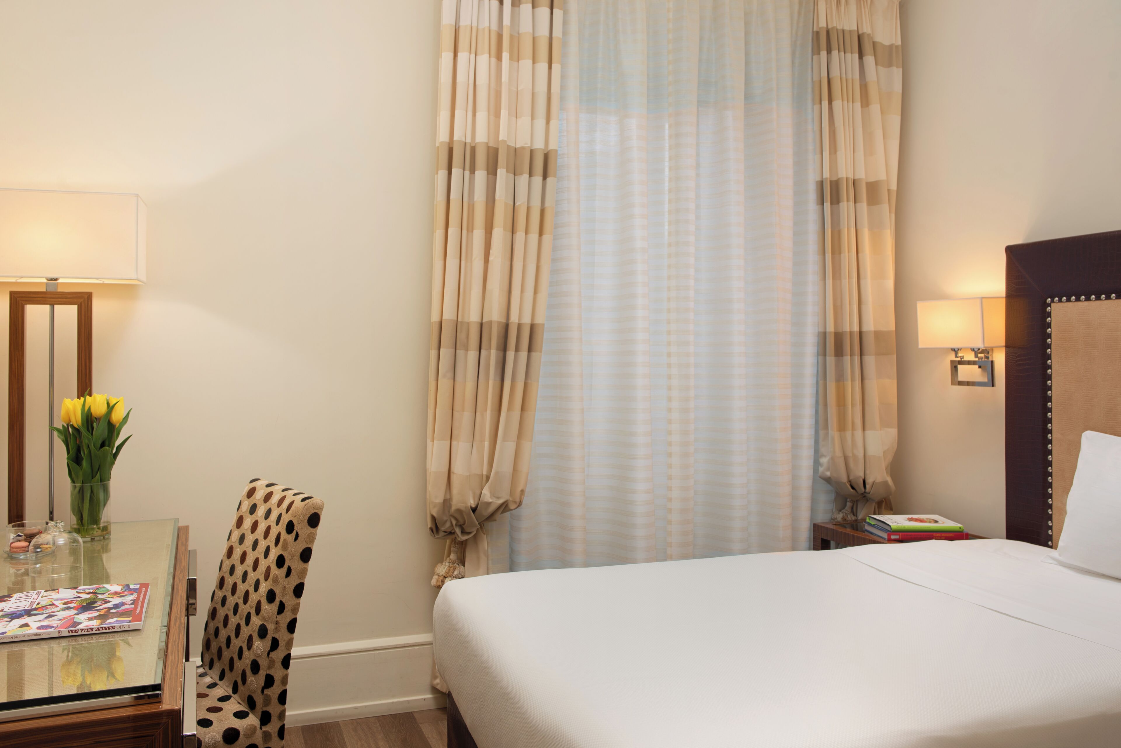 Habitación de hotel con cama, una mesita de vidrio con una revista, una silla, una lámpara y una ventana con cortinas.