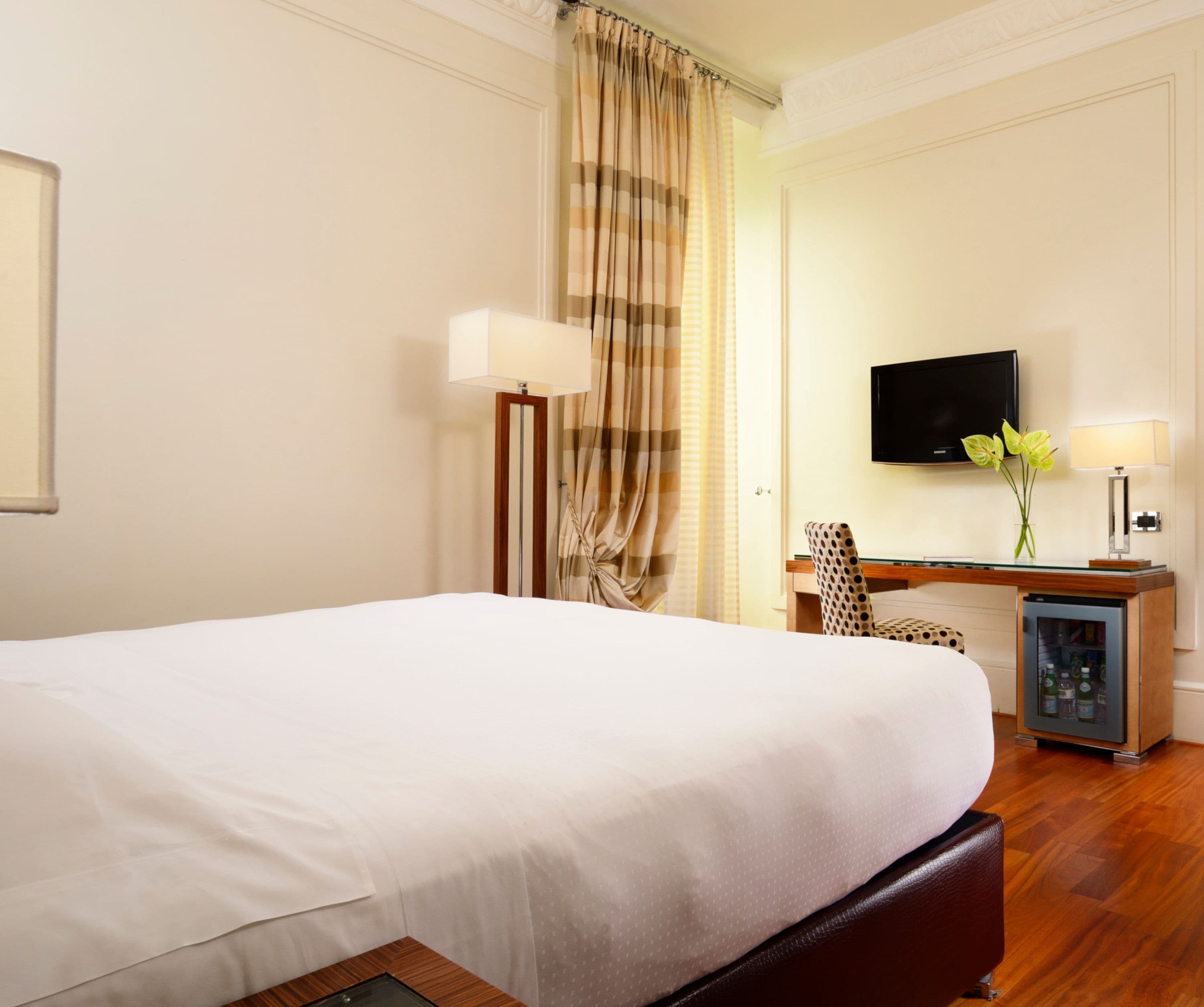 A hotel room with a large bed, a desk with a flat-screen TV, a chair, and a minibar.