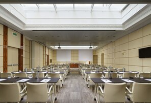 A conference room with rows of tables and chairs, a podium, and a large screen at the front.